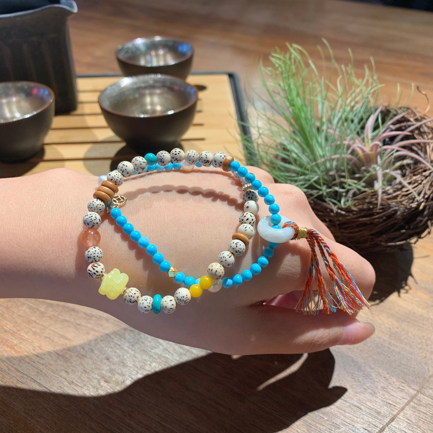 Hand wearing a stack of colorful beaded bracelets on a wooden surface with a plant in the background.